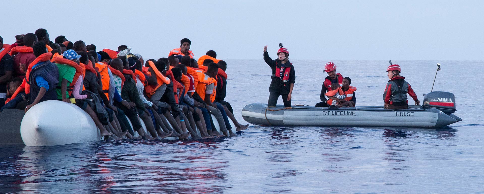 Boot mit vielen Menschen auf der linken Seite, daneben ein kleineres Motorboot mit Mission Lifeline-Crewmitgliedern, die mit Menschen auf dem zweiten Boot kommunizieren und eine Frau mit Kind an Bord haben