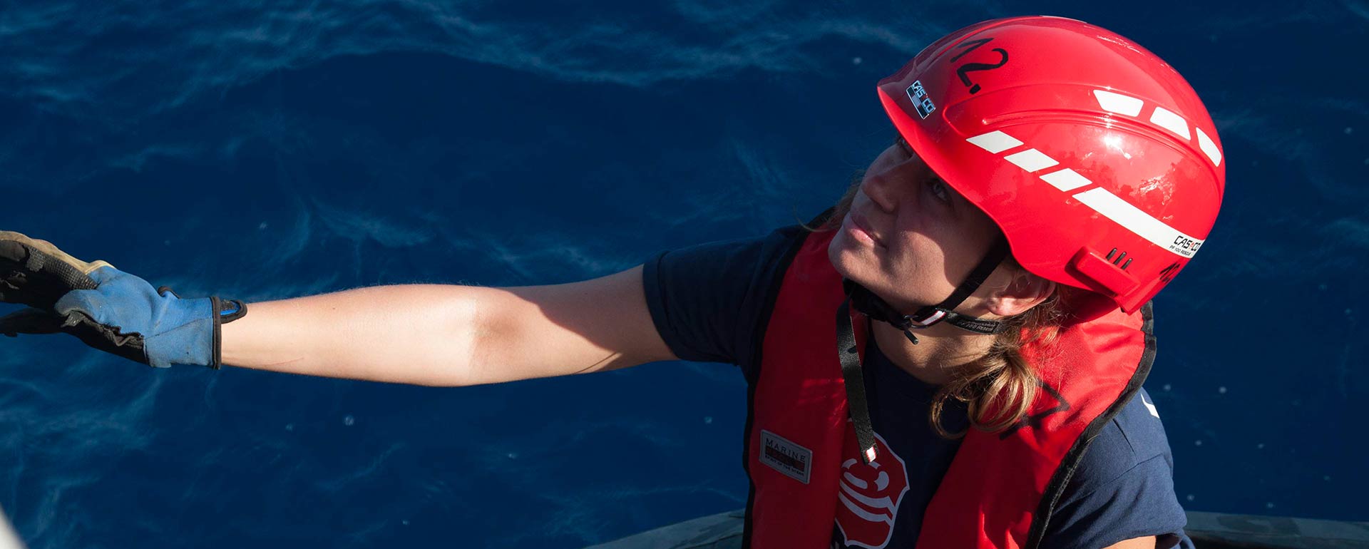 Lifeline-Crewmitglied auf Boot in Nahaufnahme, im Hintergrund das Meer, hat Helm und Handschuhe an und schaut nach oben