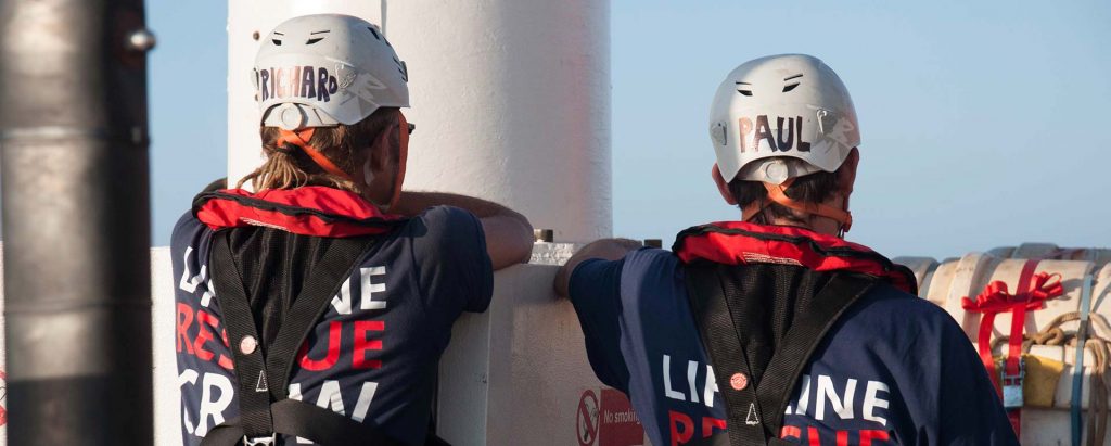 zwei Lifeline-Crewmitglieder von hinten, haben Rettungswesten an und lehnen sich an einer Säule an. Weiße Helme tragen Beschriftungen "Richard" und "Paul"