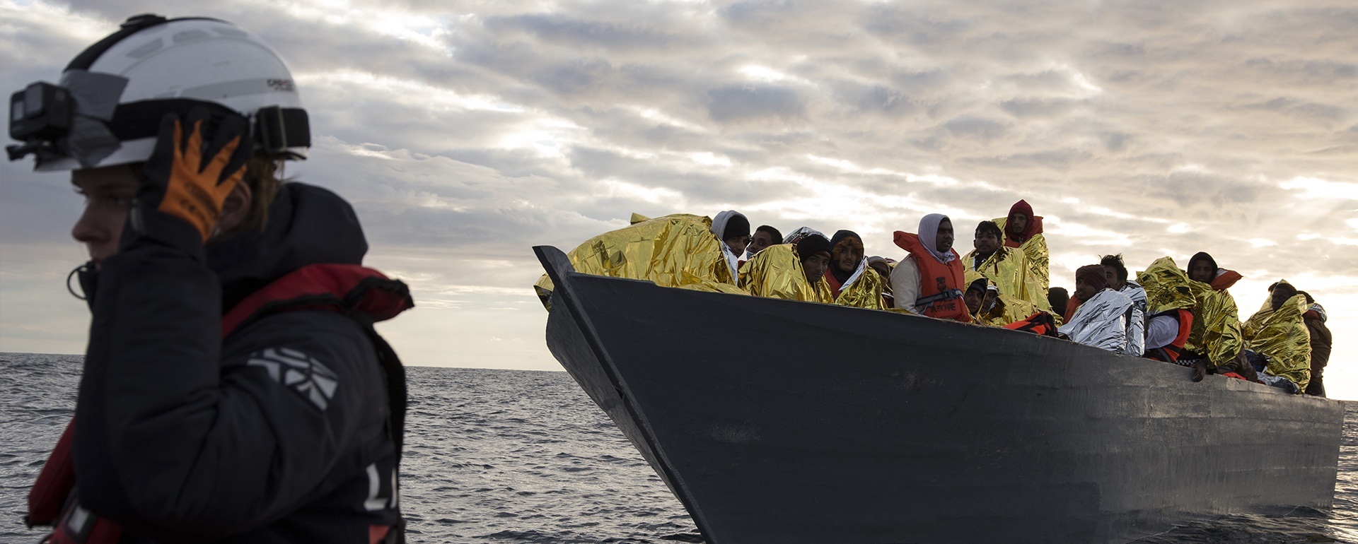 im Vordergrund Mission Lifeline Mitarbeiterin, im Hintergrund boot mit Gerettete